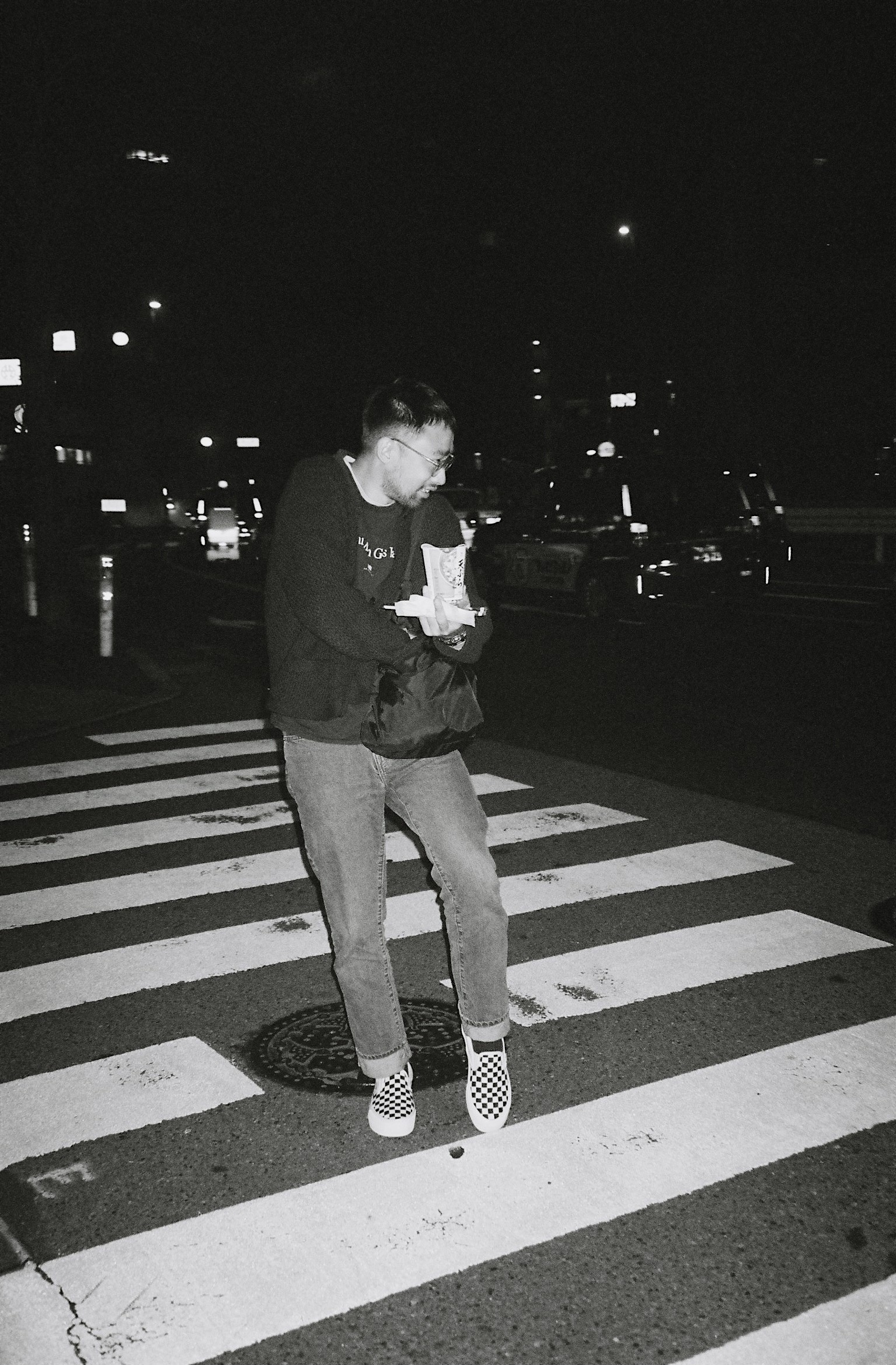A black and white picture of James Lontoc taking something out of his bag while holding cup noodles and crossing the street.